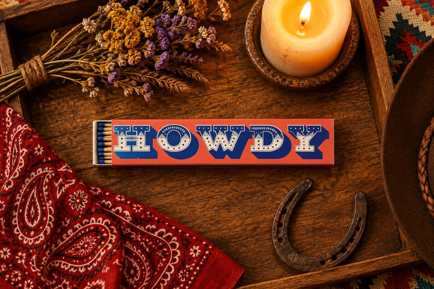 A matchbox-style novelty item with a peach-pink rectangular body, large blue and white 'HOWDY' lettering across the top, and a small stack of blue-tipped matches visible on the left side, shown on a plain white background.