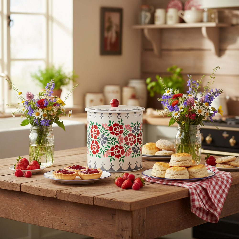 A cylindrical stainless steel biscuit barrel with a floral pattern in shades of red, green, and blue, and a white scalloped edge design around the lid, topped with a red knob.
