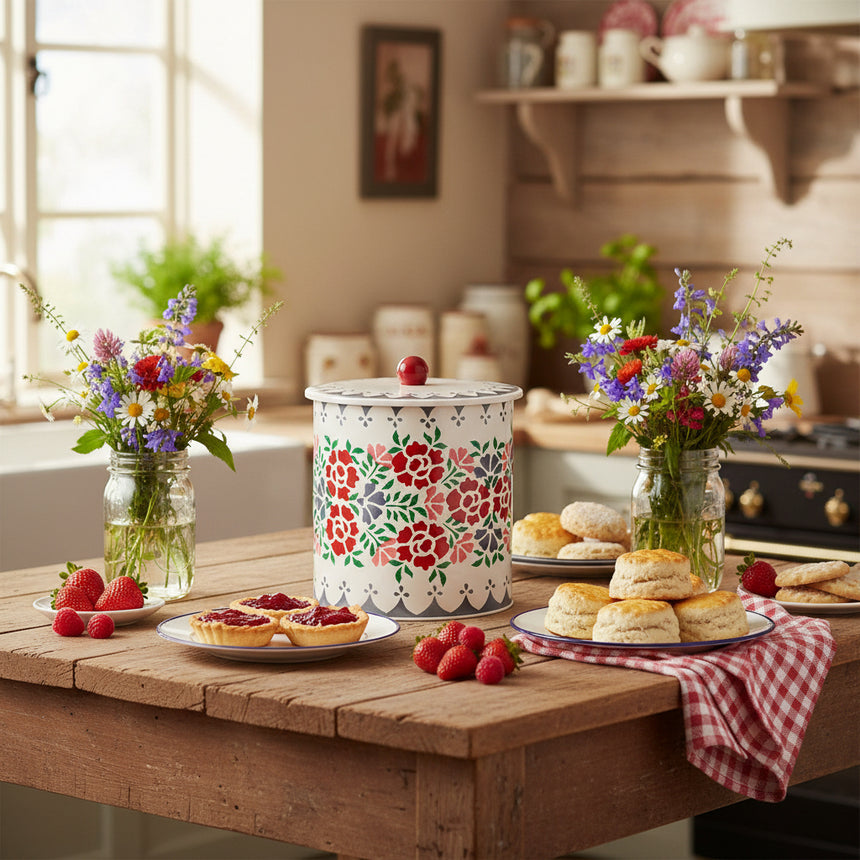 A cylindrical stainless steel biscuit barrel with a floral pattern in shades of red, green, and blue, and a white scalloped edge design around the lid, topped with a red knob.