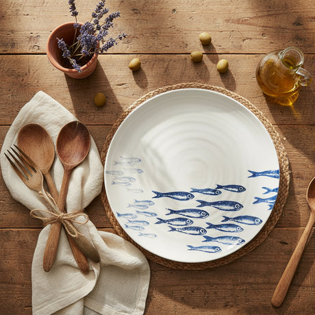 A round earthenware serving platter with hand-printed illustrations of sardines swimming in a navy blue sea, created by bliss home.