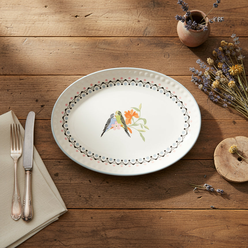 A ceramic oval serving platter with a hand-painted design of budgies and floral elements on a white background, bordered with a black pattern.
