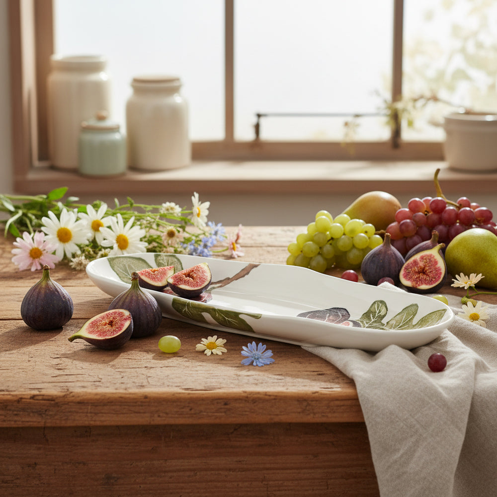 A shallow oval platter with a hand-printed illustration of fig fruits on a white background, with a leafy green border.