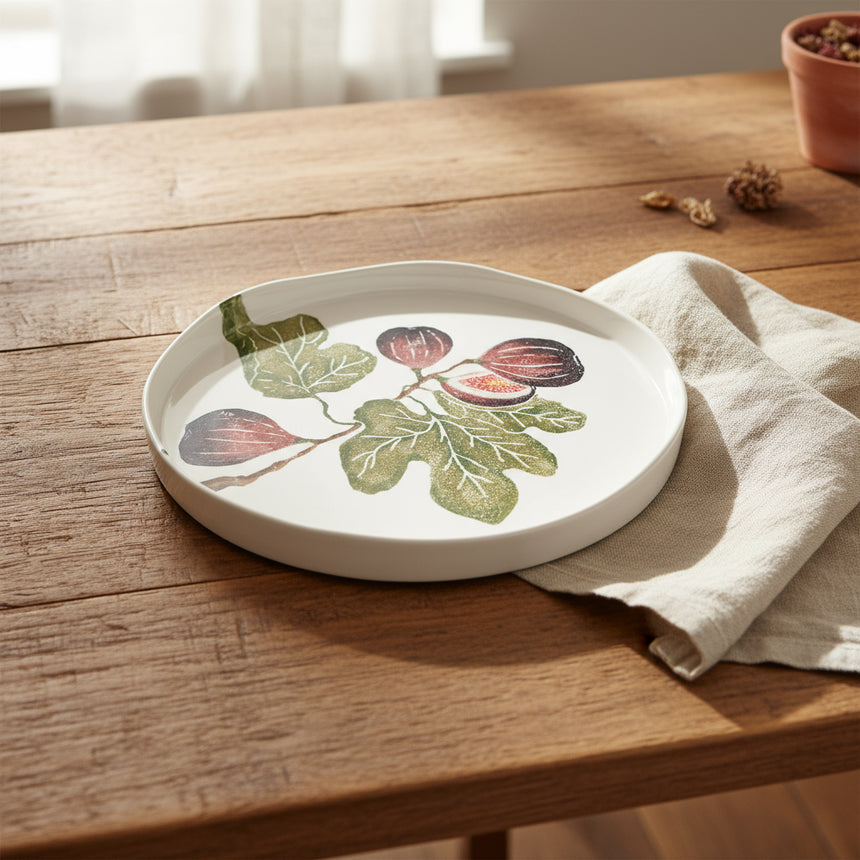 A round, white shallow serving platter with an illustration of fig fruits, leaves, and a stem, hand-printed in green and purple colors.