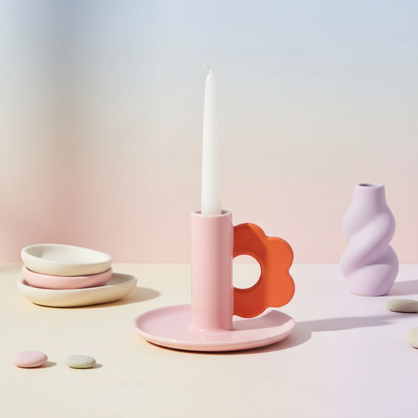 A pink stoneware candle holder with a bold orange flower loop handle on the side, designed to hold a taper candle.