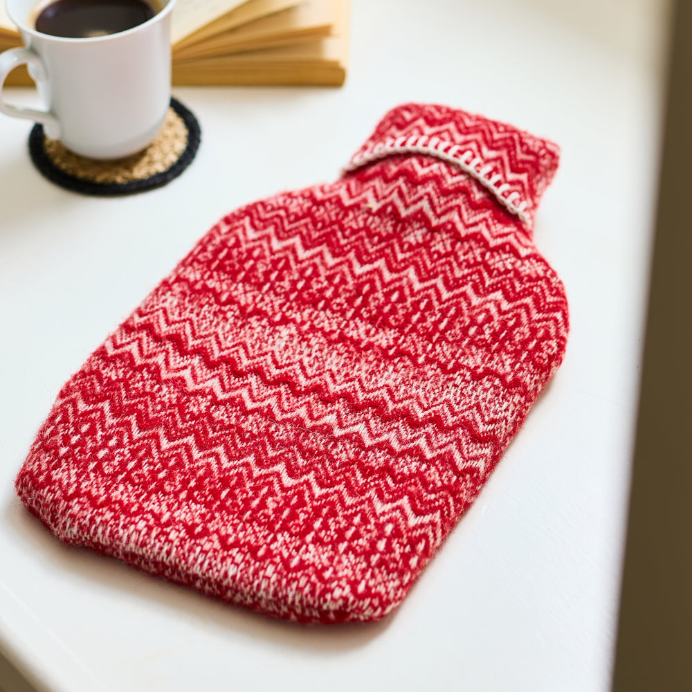 Red and white patterned hot water bottle on a table with a cup of coffee and book in the background.