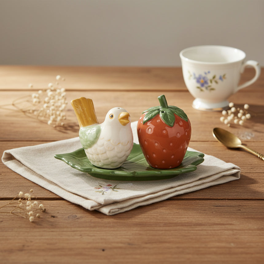 A ceramic salt & pepper featuring a small white bird with yellow and green accents and a red strawberry, both sitting on a green leaf-shaped base against a plain white background.