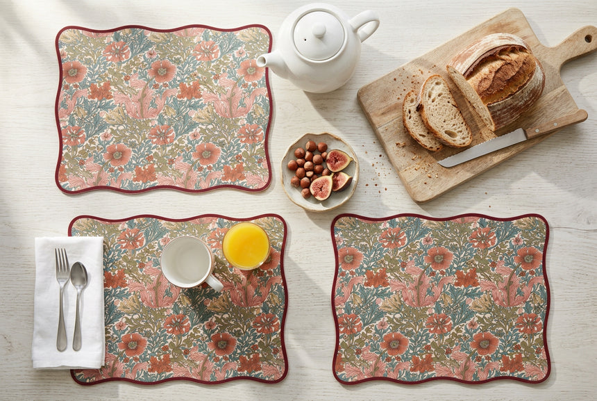 A rectangular placemat with wavy scalloped edges, featuring a dense floral and botanical pattern in muted coral, sage green, and olive tones, finished with a dark red border on a white background.