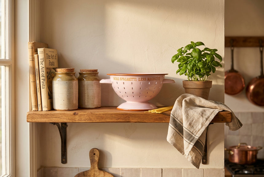 A light pink enamel colander with orange-brown trim and handles, featuring the text 'MORE SPAGHETTI LESS REGRETTI' in orange on the front.