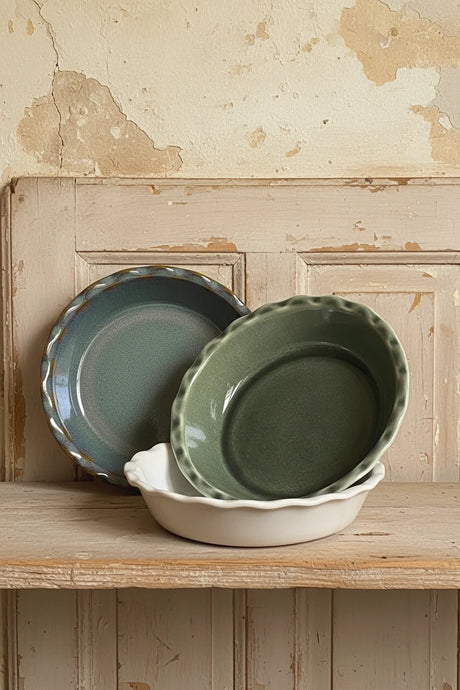 Three ceramic pie dishes  with scalloped rims displayed on a light wood surface against a plain off-white wall. The front bowl is a dark green glazed oval, the back left bowl is a muted blue-green glazed round, and the back right bowl is an unglazed cream-colored oval.