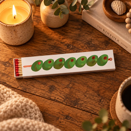 A long, narrow matchbox-style package with a cream background and a row of green olives printed across the front, each with a small red pit visible. A small bundle of red-and-tan matches sits in the match head on the left.