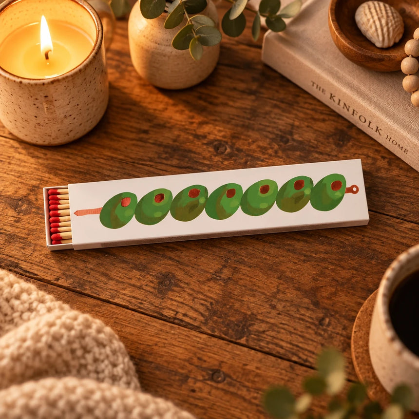 A long, narrow matchbox-style package with a cream background and a row of green olives printed across the front, each with a small red pit visible. A small bundle of red-and-tan matches sits in the match head on the left.