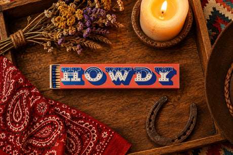 A matchbox-style novelty item with a peach-pink rectangular body, large blue and white 'HOWDY' lettering across the top, and a small stack of blue-tipped matches visible on the left side, shown on a plain white background.