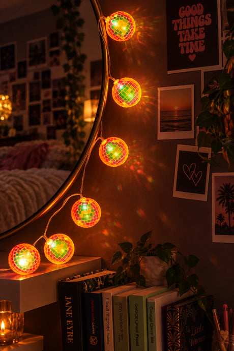 Warm white LED string lights with a mini disco ball design, glowing orange and green, hanging in a cozy room setting next to a bookshelf.