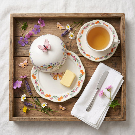 A ceramic butter dish set with a scalloped plate and matching lid, decorated with a colorful floral border, a butterfly motif on the plate, and a pink flower handle on the lid, shown on a white background.
