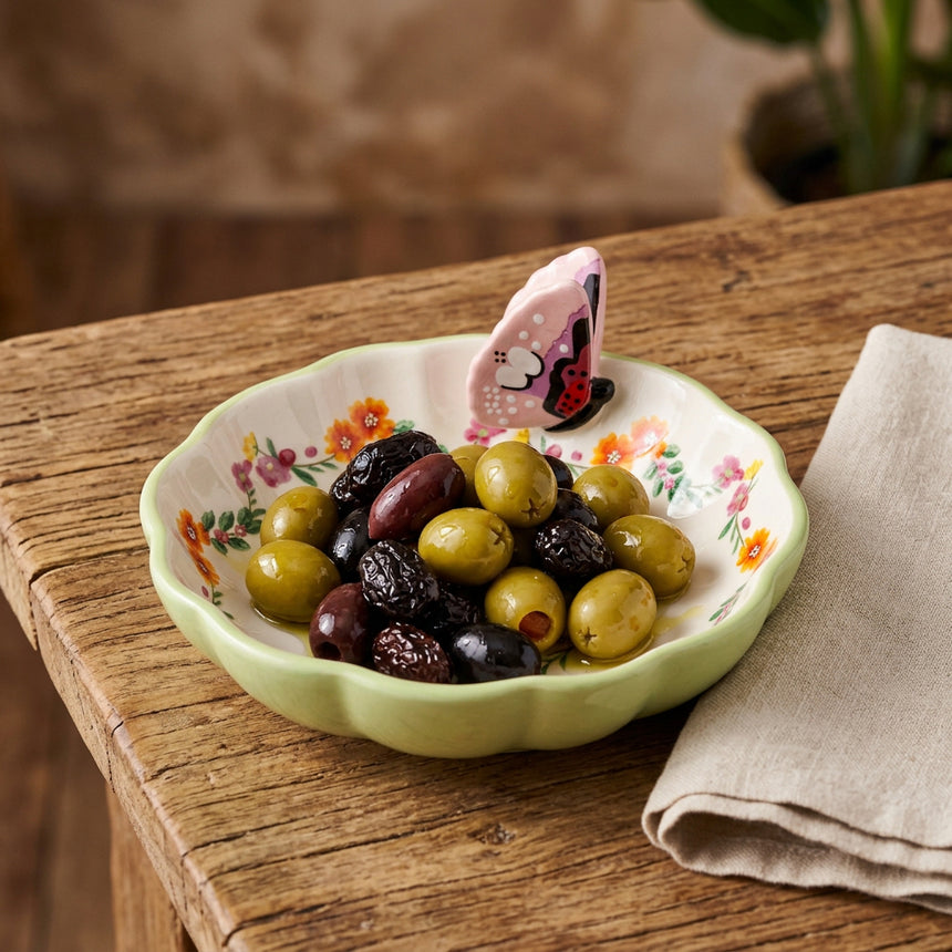 A small ceramic dish with a scalloped light green rim, a white interior decorated with orange and pink flowers, and a pink butterfly-shaped handle on the side.