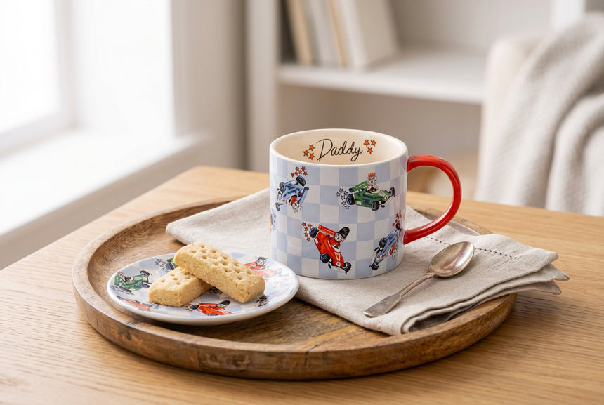A white ceramic mug with a light blue and white checkered pattern, decorated with cartoon go-karts in red, green, and blue. The inside of the mug features the word 'Daddy' in black script with small orange stars.