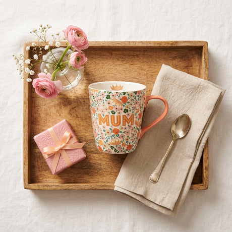 A ceramic mug with a white background covered in a dense floral pattern of orange, green, and pink flowers. The word 'MUM' is printed in orange across the center, and a small gold crown is visible on the inside of the mug near the rim.