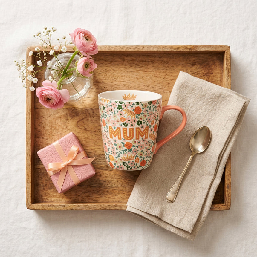 A ceramic mug with a white background covered in a dense floral pattern of orange, green, and pink flowers. The word 'MUM' is printed in orange across the center, and a small gold crown is visible on the inside of the mug near the rim.