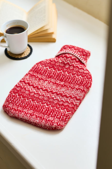 Red and white patterned hot water bottle on a table with a cup of coffee and book in the background.