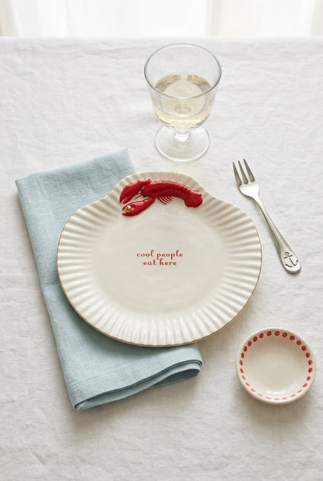 A scallop-shaped  plate in off-white with a red lobster illustration and the words 'cool people eat here' printed in red, shown on a light blue background.