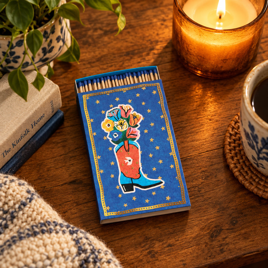 A decorative matchbox with a blue background, gold star border, and an illustration of a cowboy boot filled with colorful flowers. The top edge shows several wooden matches with blue tips.