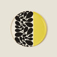 Round ceramic plate with black leaf pattern on one half and yellow on the other, on a beige background.