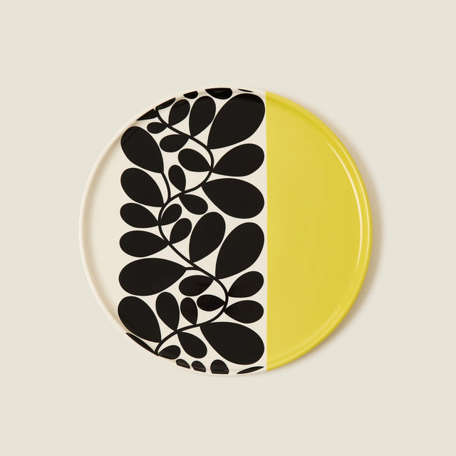 Round ceramic plate with black leaf pattern on one half and yellow on the other, on a beige background.