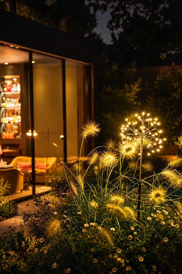 A garden scene at night with glowing dandelion-style lights among tall grasses in front of a lit building with large glass windows.