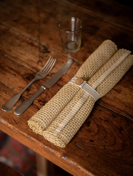 Rolled fabric placemats  with a wooden button on a wooden table with cutlery and a glass.