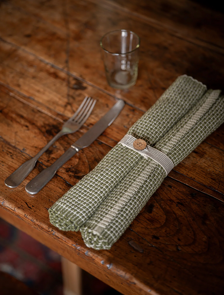 Rolled green fabric placemats  with a button on a wooden table with cutlery and a glass.
