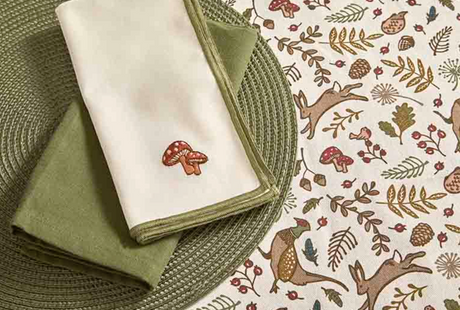 A folded white napkin with a small embroidered red-and-white mushroom sits on top of an olive green folded napkin, placed on a woven green placemat over a cream tablecloth printed with woodland deer, leaves, and mushrooms.