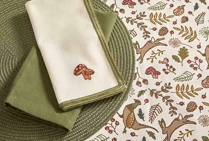 A folded white napkin with a small embroidered red-and-white mushroom sits on top of an olive green folded napkin, placed on a woven green placemat over a cream tablecloth printed with woodland deer, leaves, and mushrooms.