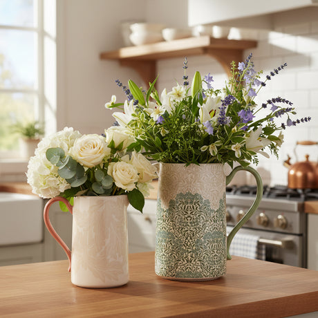Two ceramic pitchers on a white background, one small with a coral handle and one larger with a green handle and a blue-green floral pattern over an off-white base.