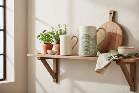 Two ceramic pitchers on a white background, one small with a coral handle and one larger with a green handle and a blue-green floral pattern over an off-white base.