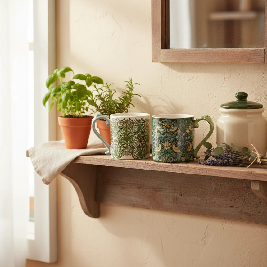 Two ceramic mugs with ornate handles, featuring a William Morris-style floral and bird pattern in green, cream, and yellow tones, shown on a white background.