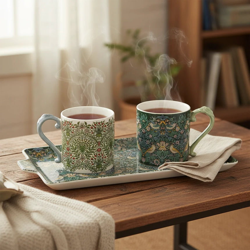 Two ceramic mugs with ornate handles, featuring a William Morris-style floral and bird pattern in green, cream, and yellow tones, shown on a white background.