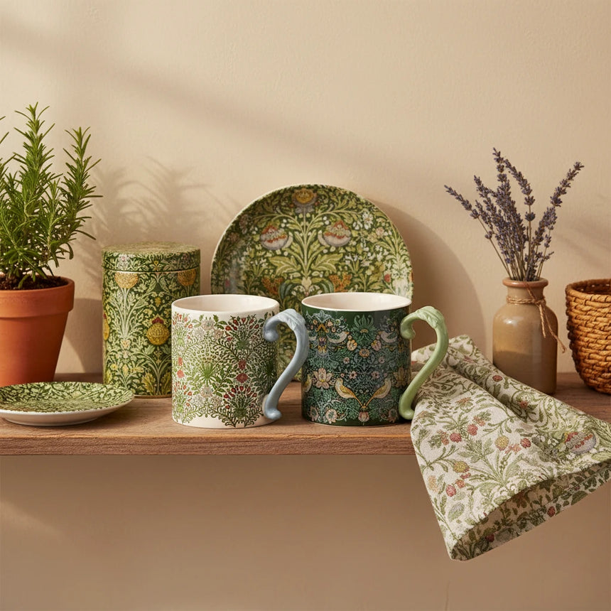 Two ceramic mugs with ornate floral and bird patterns, shown side by side on a plain white background. One mug has a light blue handle and a cream base with green and red botanical motifs.