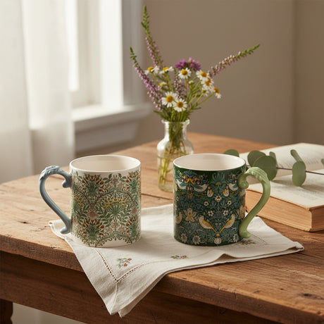 Two ceramic mugs with ornate floral and bird patterns, shown side by side on a plain white background. One mug has a light blue handle and the other has a pale green handle.