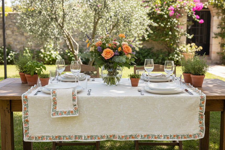 A folded beige cotton tablecloth with a subtle damask pattern and a decorative border featuring orange flowers and green leaves, shown on a solid white background.