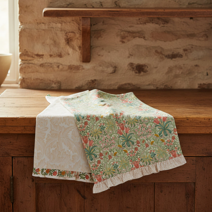 Two fabric tea towels with botanical patterns, one featuring a light beige damask design and the other a green, orange, and beige floral pattern with a ruffled edge.