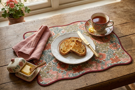 A rectangular placemat with wavy scalloped edges, featuring a dense floral and botanical pattern in muted coral, sage green, and olive tones, finished with a dark red border on a white background.