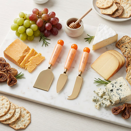 Three cheese knives with peach-colored handles and orange ball tips, displayed diagonally on a solid white background.