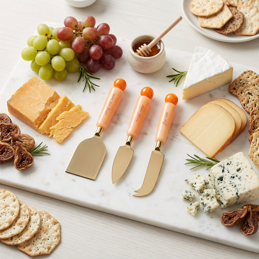 Three cheese knives with peach-colored handles and orange ball tips, displayed diagonally on a solid white background.