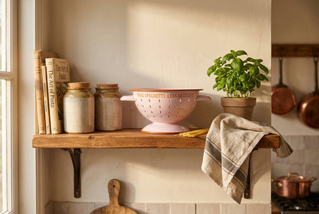 A light pink enamel colander with orange-brown trim and handles, featuring the text 'MORE SPAGHETTI LESS REGRETTI' in orange on the front.