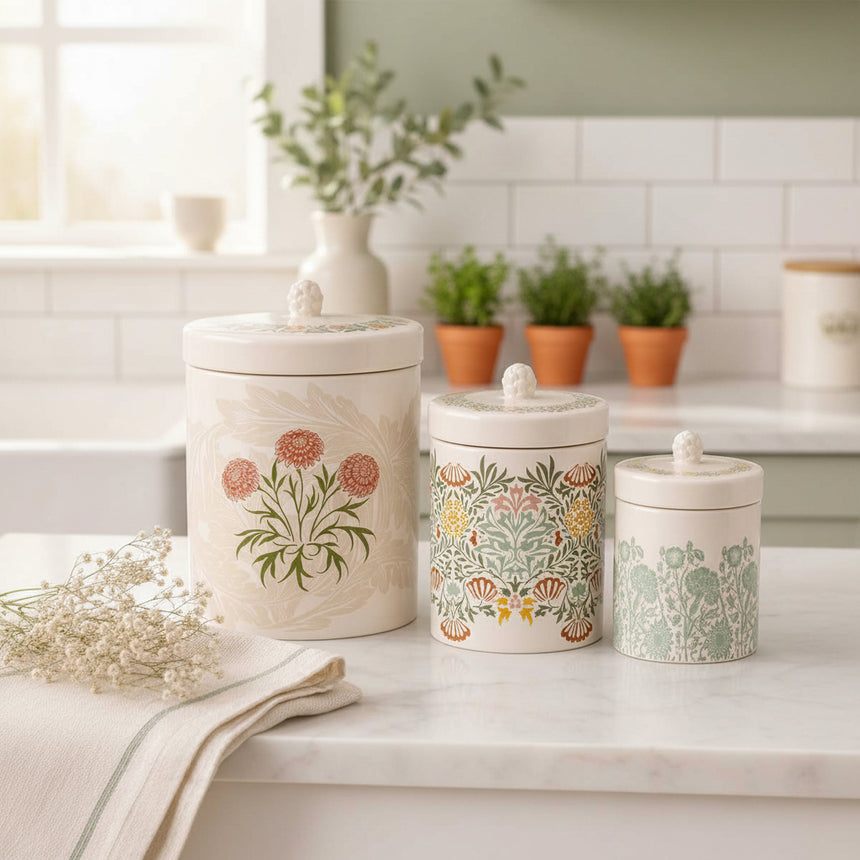 Three ceramic canisters with matching lids are arranged on a white marble surface against a white paneled wall. The canisters have a warm cream base with floral and botanical patterns in muted green, peach, orange, and beige tones.